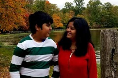 一名身穿绿白条纹衬衫的印度裔少年，看着站在五颜六色的秋天树前、身穿红色衬衫的母亲