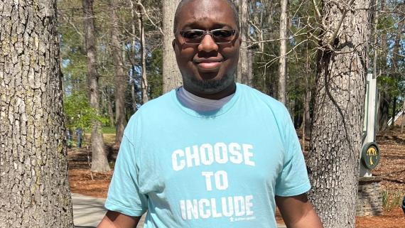 戴眼镜的年轻黑人男子站在一件蓝绿色t恤上，上面用白字写着“选择包括”