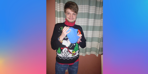 一个年轻人穿着一件黑色毛衣，上面有一棵圣诞树，上面有史努比和伍德斯托克。里面穿件红衬衫。他拿着玩具收音机，对着镜头微笑